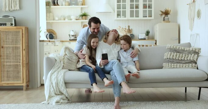 Happy blogger parents and kids hugging on home sofa, holding mobile phone, taking family self video, speaking on conference call, watching online content, smiling, laughing