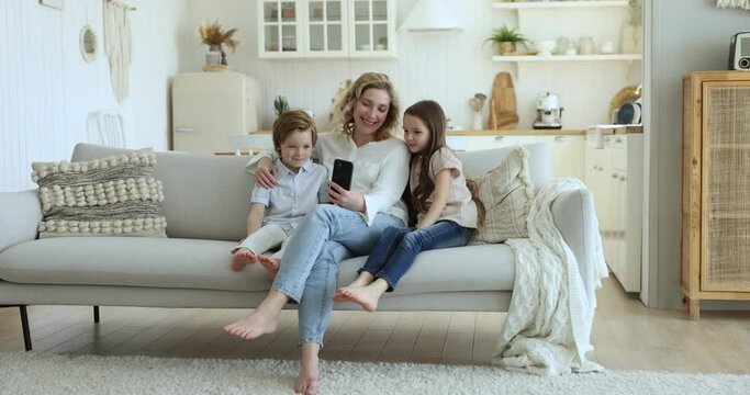 Happy family blogger mom and two kids taking self video on home couch, holding smartphone, hugging children, resting in living room, making video call, enjoying online communication