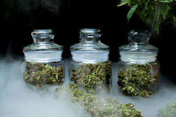 The marijuana buds in a clear glass jar sit on a smoky floor at the black background