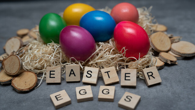 Colorful eggs lie in a straw nest with wooden decoration around. On the table are letters that spell out the word "Easter Eggs". View from above.