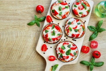 Mini eggplant pizzas-eggplants with pizza sauce,olive oil,tomatoes,mozzarella cheeses,basil and peppers on wooden pizza peel with wooden background.Healthy summer vegetarian food.Top view.Copy space