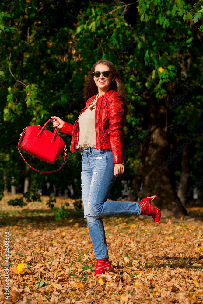 Poster Young woman in fashion red jacket and blue jeans in autumn park