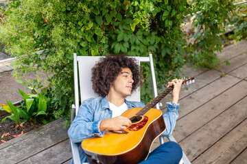  Beautiful young Afro-Italian mixed race man gen Z Man in casual wear playing guitar in the garden. A multiracial boy plays a musical instrument rests and relaxes outdoors. Spending time at home