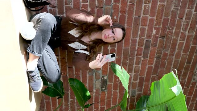 beautiful teenage girl on background of brick wall paints her eyelashes looks into reflection of phone flowerpot black mascara brown blouse with rhombuses modern fashionable outfit is preparing to go
