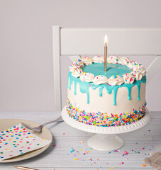 Birthday party with vanilla buttercream Cake, lit gold candle, blue drip and sprinkles on a light grey white background - 582512574
