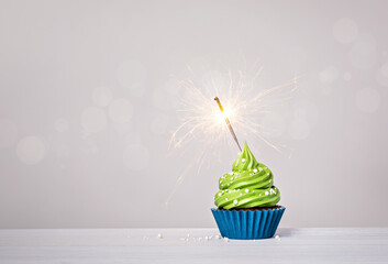 Green cupcake with sprinkles and lit sparkler on a white grey background. - 582512939