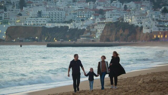 Telephoto shot - a family of four, including a 40-year-old father, mother, 12-year-old son, and 5-year-old daughter, stroll along the ocean coastline in Portugal holding hands, with the subdued