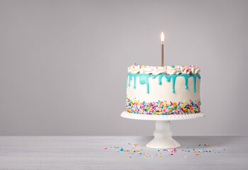 Vanilla Birthday Cake with a lit gold candle, teal blue drip and colorful sprinkles on a light grey white background