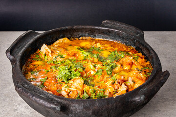 Delicious Brazilian fish moqueca with tomato, onion, olive oil, coriander and orocum seed. Made in clay pot on white table and black background