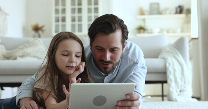 Millennial caring dad and cute little daughter girl resting on floor at home, using digital tablet, shopping, watching movie, smiling. Father and child enjoying leisure, reading book on Internet