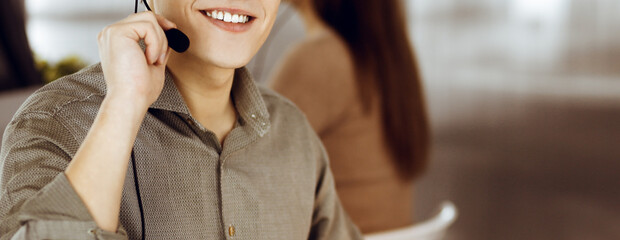 Young dark-haired guy in a green shirt and headsets is talking to a client, while sitting at the desk, working together with a female colleague in a modern office. Call center operators at work