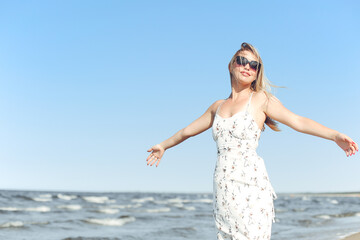 Fototapeta na wymiar Happy blonde beautiful woman on the ocean beach standing in a white summer dress and sun glasses, open arms