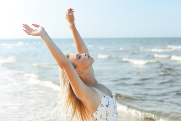 Fototapeta Happy blonde beautiful woman on the ocean beach standing in a white summer dress, raising hands obraz