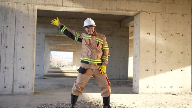 cheerful positive male builder in a white hard hat and uniform is dancing on the background of construction