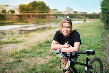 Obraz premium A woman likes active recreation, often rides bicycles together with her husband or friends, now during another ride she rests by the river in the city and smiles