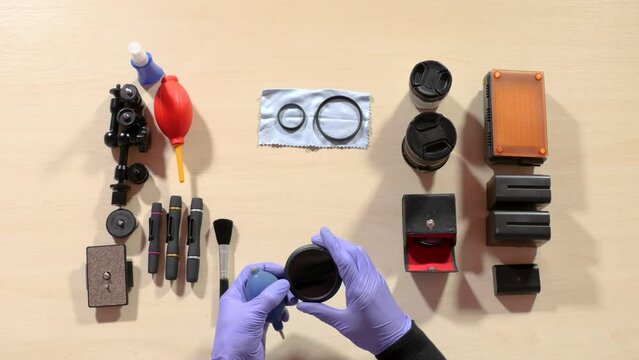 Professional serviceman in blue gloves cleans the lens glass with a brush on the table with a set of photographic accessories, batteries, cleaners, napkins.