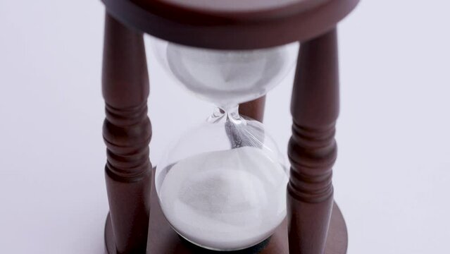Antique sand watch isolated on white background, two glass bulbs in three legged wooden stand, white sand trickle down on pile, measuring time. Close up, high angle, 4K Ultra HD.
