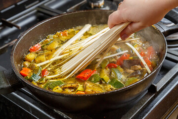 A woman's hand puts udon in a pan with stewed vegetables.