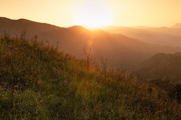 Breathtaking mountain landscape - bright golden sunset with sun,  sunbeams, rays, glare above lush...