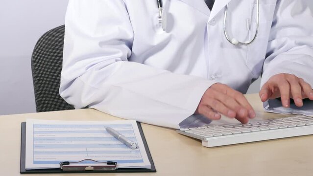 Male in white coat with stethoscope on neck sitting at table, typing on computer keyboard, medical record, silver pen, health care, work in hospital. Close up, 4K Ultra HD.
