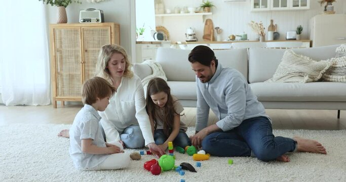 Loving parents helping kids to build toy tower on floor, stacking colorful construction blocks on white soft carpet, playing family learning games on warm floor, enjoying home activity