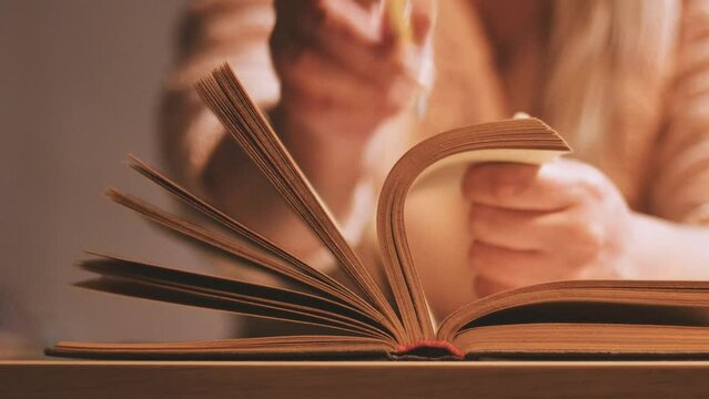 A young unrecognizable girl at the table puts a pen between the pages of a book, stops reading, learning, studying, and education process, and takes a break to rest and relax.