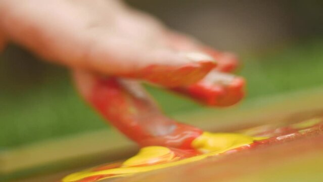 Close-up shot of a finger smearing liquid drops of bright saturated yellow and red acrylic paint on the surface of an abstract background.