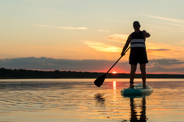 A man in shorts on a sapboard with an oar swims in the lake against the backdrop of the sunset sky.