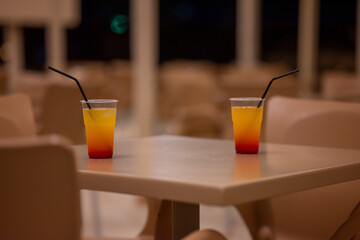Two Plastic Glasses With Cold Cocktail Sunrise With Black Cocktail Straw on Plastic Table in Hotel Restaurant At Night Moraitika, Corfu, Greece.