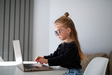 beautiful, stylish, fair-haired girl of terh years, studying at a computer, a girl learns English, studies online with a teacher, a girl sits on a chair at a white table, a child with glasses