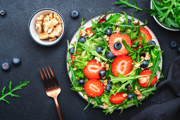 Strawberry and herbs healthy salad with arugula, blueberries and walnuts, black kitchen table, place for text. Fresh useful vegan dish for healthy eating