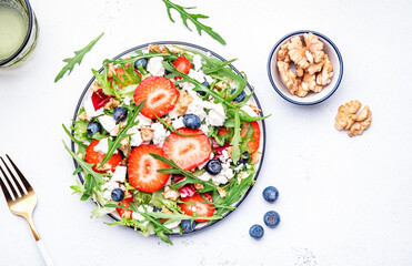 Strawberry and herbs healthy salad with arugula, blueberries, soft white feta cheese and walnuts, white kitchen table, place for text. Fresh useful dish for healthy eating