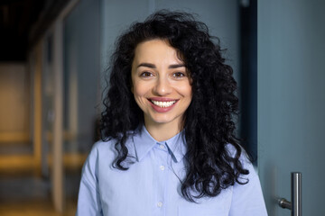 Close-up portrait of mature adult business woman, boss smiling and looking at camera, pleased hispanic woman with curly hair in blue shirt inside office at workplace in hall corridor.