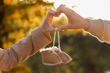 Man and pregnant wife are doing heart gesture with hands and holding warm baby shoes on nature...
