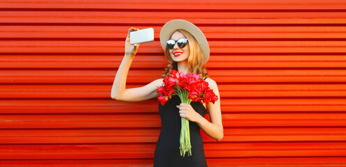 Portrait of beautiful young woman taking selfie with smartphone holding bouquet of pink flowers in summer straw round hat on red background - Powered by Adobe