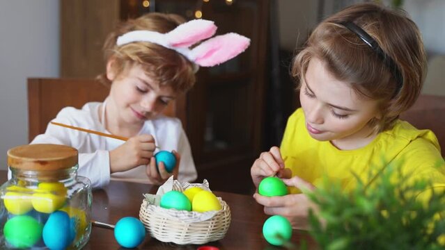Easter Family traditions. Two caucasian happy school children kids with bunny ears paint and decorate eggs with paints for holidays while sitting together at home table. Kids having fun together