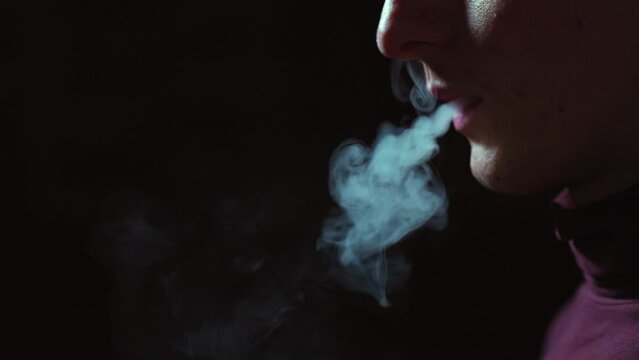 Electric device for smoking. A man smokes an electronic cigarette in the dark. Vape. E-cig, vsping. Concept: the harm of smoking. Slow motion, smoke on a dark background, A man without a face holds an