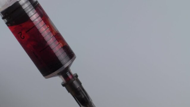 Extreme close-up drawing in blood into syringe. Macro shot medical procedure syringe pulling red liquid into saline solution isolated on gray background