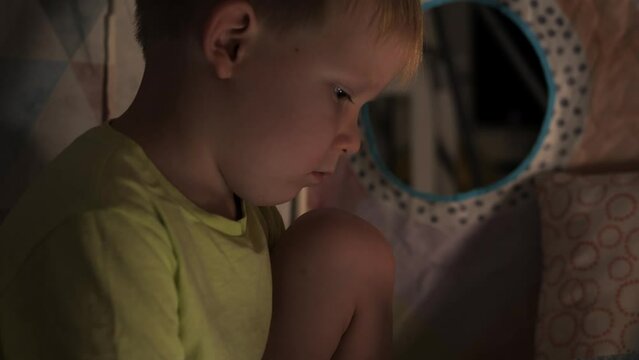 A little boy of 5 years old is sitting alone in a children's tent in the evening and is bored, looking at a book. Side view. High quality 4k footage