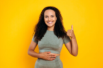 Happy brazilian young woman in sportswear, keeps one hand on her stomach, the other one shows thumb...
