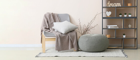 Chair with pillows, knitted plaid, pouf and shelving unit near light wall in room