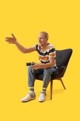 Handsome man playing video game in black armchair on yellow background
