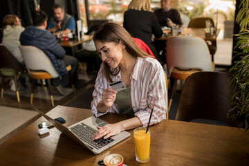 A young woman sitting at a coffee shop makes an online purchase using her credit card and laptop