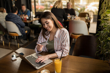 A young woman using her credit card and laptop for online shopping while sitting in a restaurant 