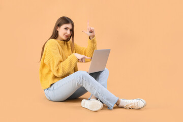 Young woman with laptop showing loser gesture on beige background