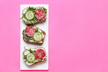 Plate of tasty sandwiches with cream cheese, salami, cucumber and arugula on color background
