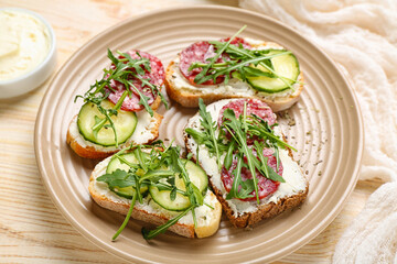 Plate of tasty sandwiches with cream cheese, cucumber, arugula and salami on light wooden table, closeup