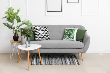 Cozy sofa with cushions and houseplants on tables near white wall