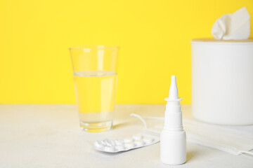 Nasal drops with pills, medical mask and glass of water on table near yellow wall. Allergy concept