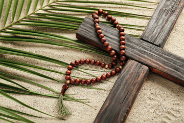 Wooden cross with prayer beads and palm leaf on sand, closeup. Good Friday concept